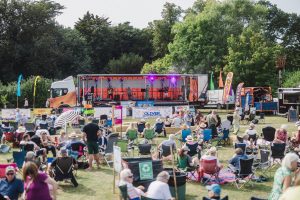 A band plays from a trailer of a lorry, entertaining a field of people sitting on picnic chairs