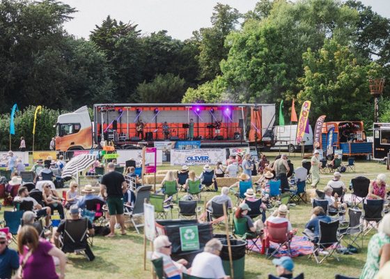 A band plays from a trailer of a lorry, entertaining a field of people sitting on picnic chairs