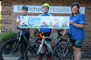 Cyclists outside the Hospice building holding a sign promoting the St Nic's Cycle Ride