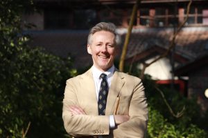 A man smiling stood in front of some trees. He has his arms crossed.