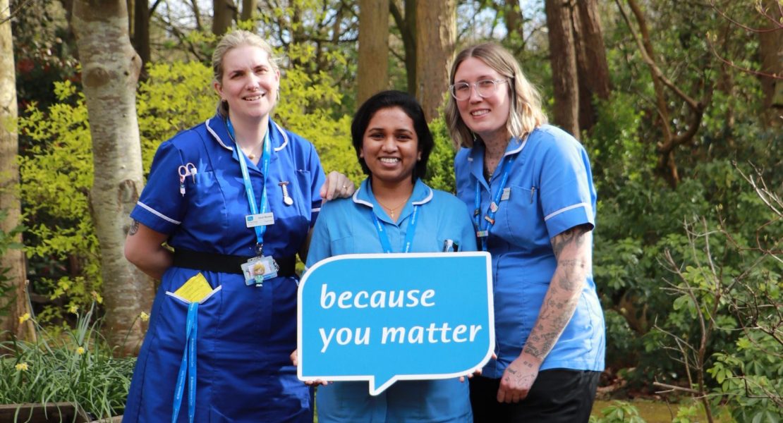Three Hospice clinical staff in uniform standing in the Hospice garden holding the logo speech bubble with the words 'Because you matter'