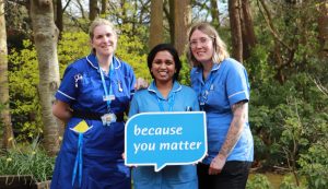 Three Hospice clinical staff in uniform standing in the Hospice garden holding the logo speech bubble with the words 'Because you matter'