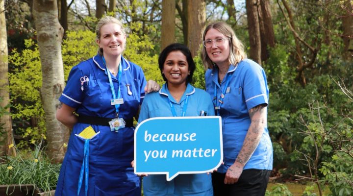 Three Hospice clinical staff in uniform standing in the Hospice garden holding the logo speech bubble with the words 'Because you matter'