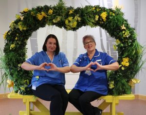 two hospice nurses posing in a flower wreath with hands in the shape of hearts