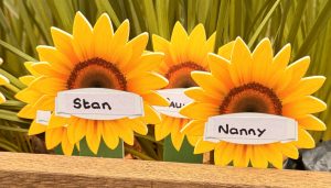 Sunflower memorial markers labelled “Stan” and “Nanny” placed in a wooden garden bed with green plants behind.