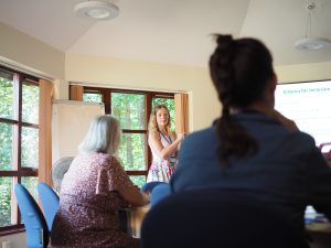 Trainer delivering an educational presentation to attendees in a meeting room with a projected slide and flip chart.