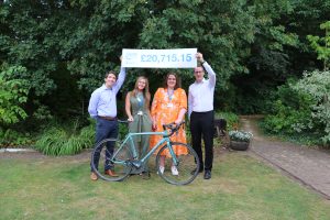 Four people stand in a garden beside a bicycle, holding a large sign showing the St Nicholas Hospice Care Cycle Ride total of £20,715.15. Trees and landscaped greenery form the background, with the fundraising total displayed prominently above the group.